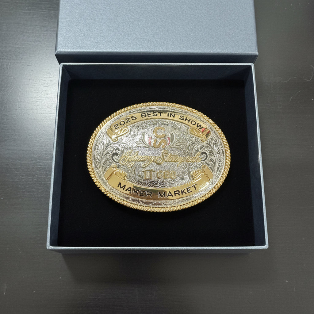 A large oval silver-tone belt buckle with gold-tone trim and raised decorative text, displayed inside an open black presentation box on a desk.